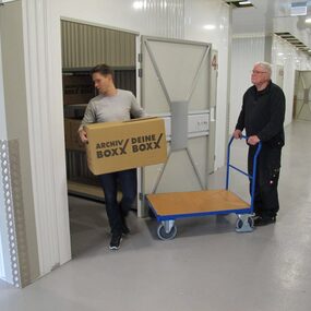Zwei Männer lagern Kartons in einem modernen Selfstorage-Lagerraum mit Transportwagen ein.